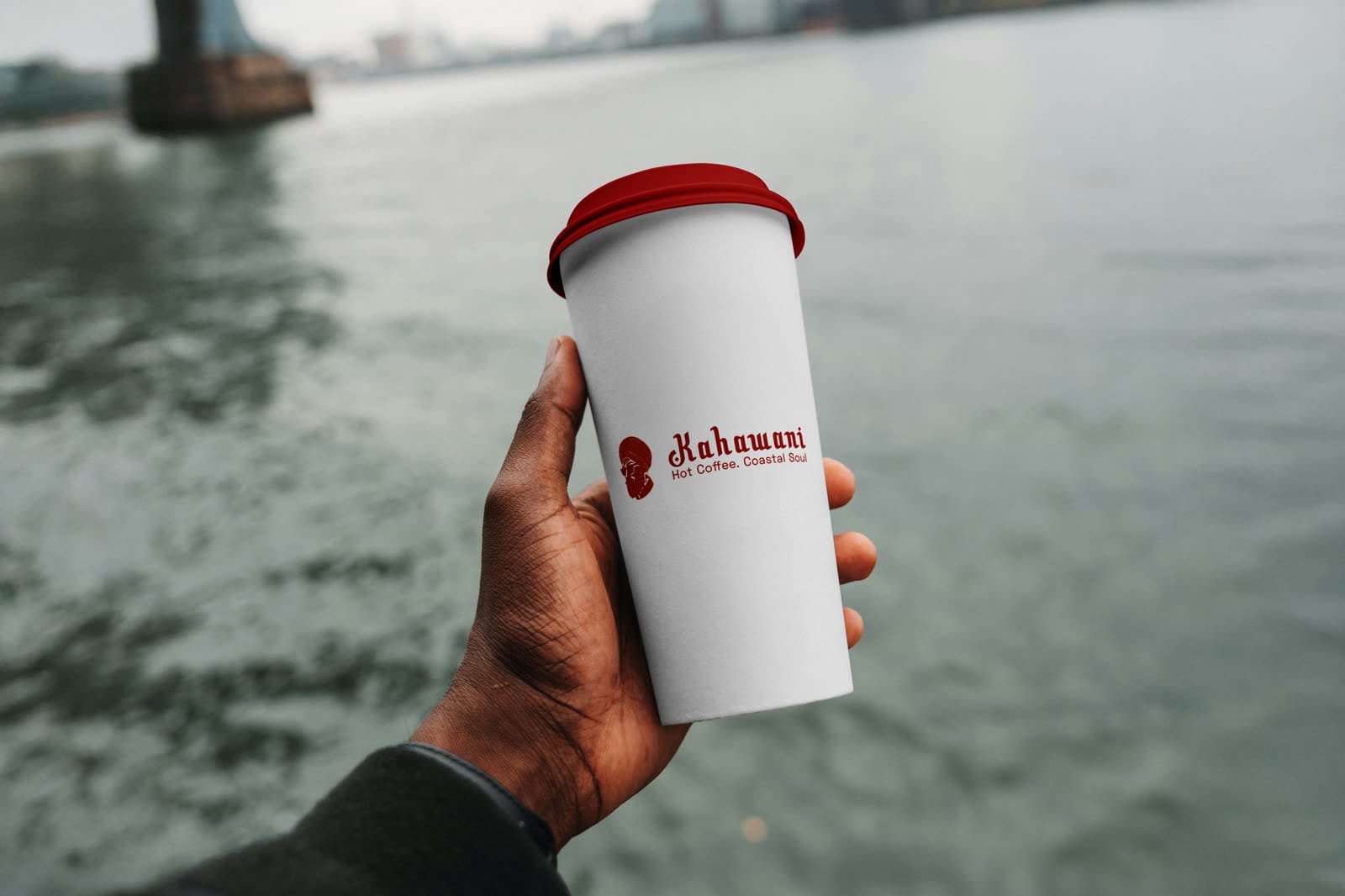 Hand holding a white Kahawani takeaway cup with a red lid, portrait mark and tagline, against a coastal waterfront.