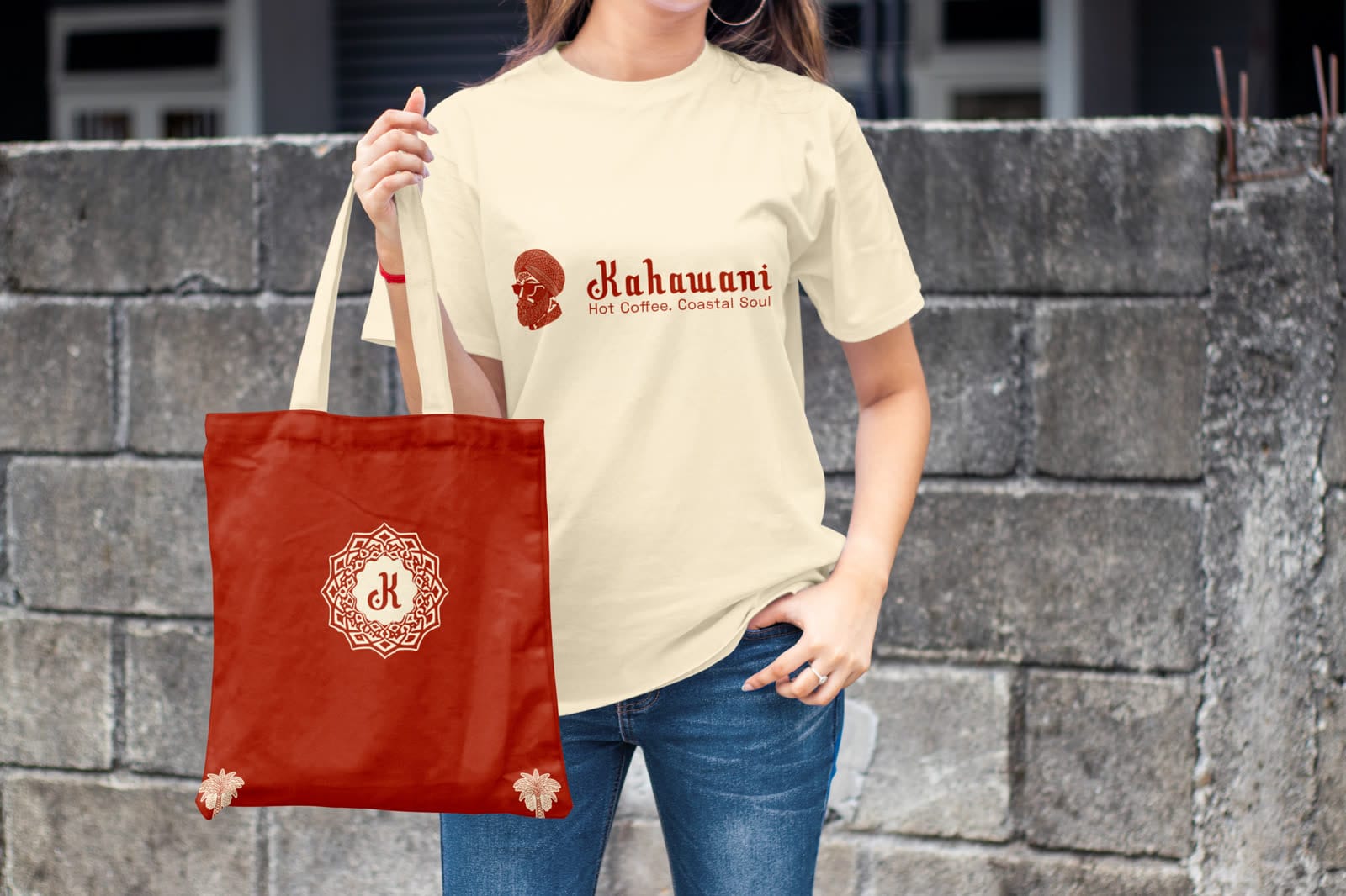 Cream Kahawani t-shirt with the turban portrait and tagline, held with a red tote printed with an ornamental K monogram.